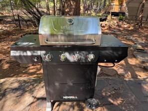Een NEXGRILL barbecue met vier branders en een digitale thermometer, buiten geplaatst met bomen en een houten constructie op de achtergrond.