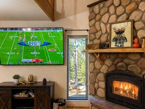 Una sala de estar con chimenea de piedra, un televisor montado mostrando un partido de fútbol y un ventanal con vistas a un bosque.