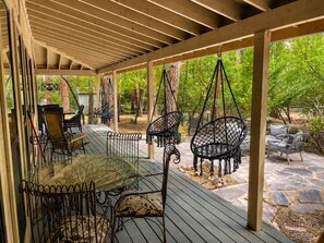 An outdoor covered patio area with a glass table and metal chairs, surrounded by stone flooring and greenery.