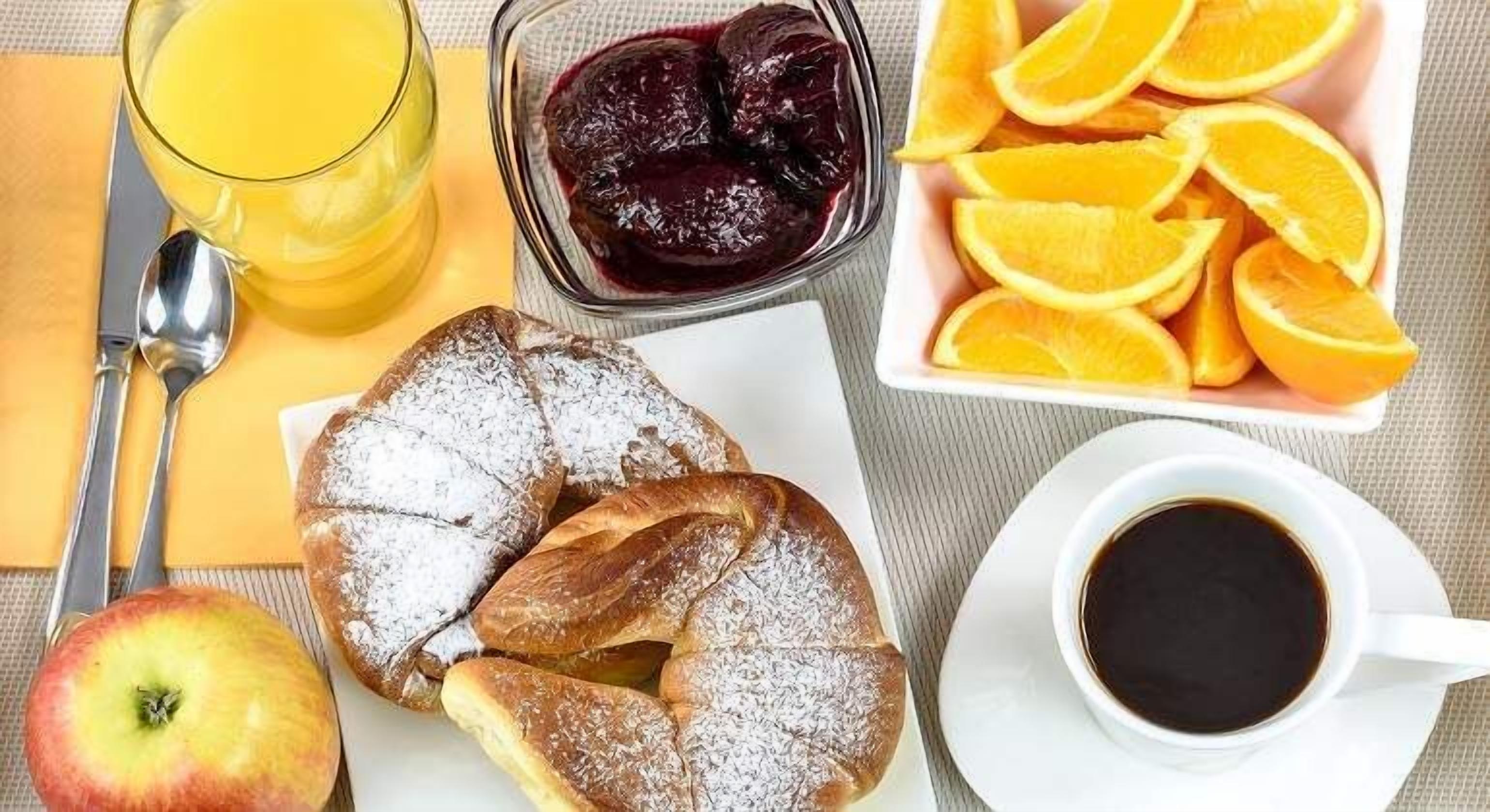 Un desayuno con un vaso de zumo de naranja, un bol de compota de frutas, rodajas de naranja, un pastel, una manzana y una taza de café.