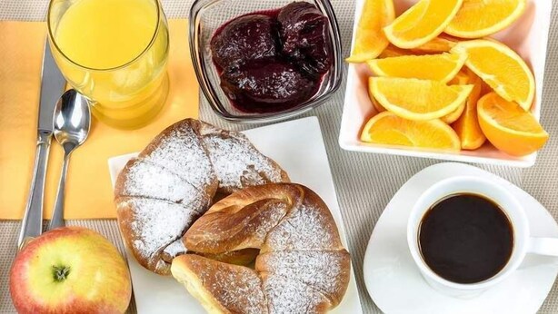 Disposição para o café da manhã com um copo de suco de laranja, uma tigela de compota de frutas, fatias de laranja, um pastel, uma maçã e uma xícara de café.