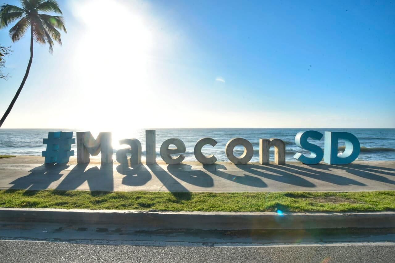Een bord met de tekst 'Malecon SD' voor een achtergrond van de oceaan en een palmboom.
