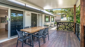 A covered outdoor dining area with a wooden table and chairs, a sliding glass door leading to a kitchen, and a view of a residential area with greenery.