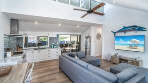 A spacious living area with a large grey sofa, a dining table with chairs, a kitchen with white cabinets, and a ceiling fan. - Castaway Cottage - Rainbow Beach (Rainbow Beach)