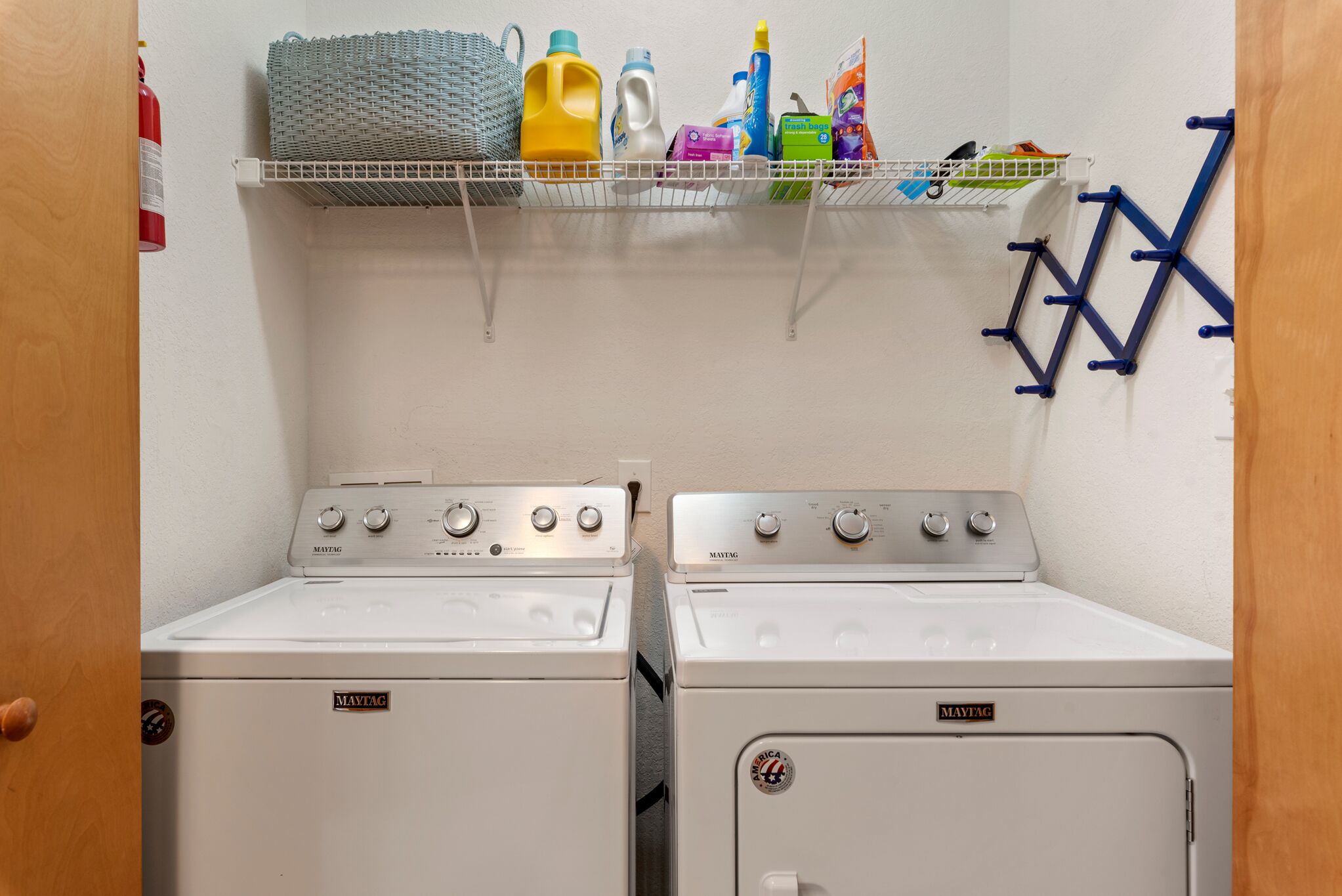 A laundry room with two Maytag dryers, a shelf with cleaning supplies, and a blue drying rack.