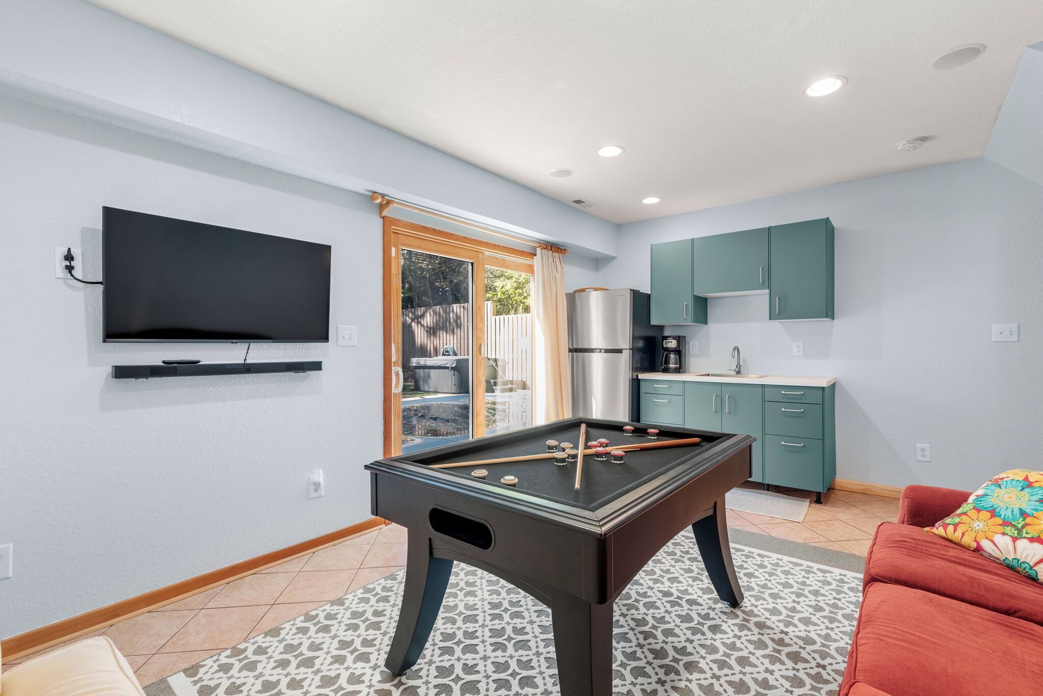 A modern living room with a pool table, a television mounted on the wall, and a kitchen area with teal cabinets.