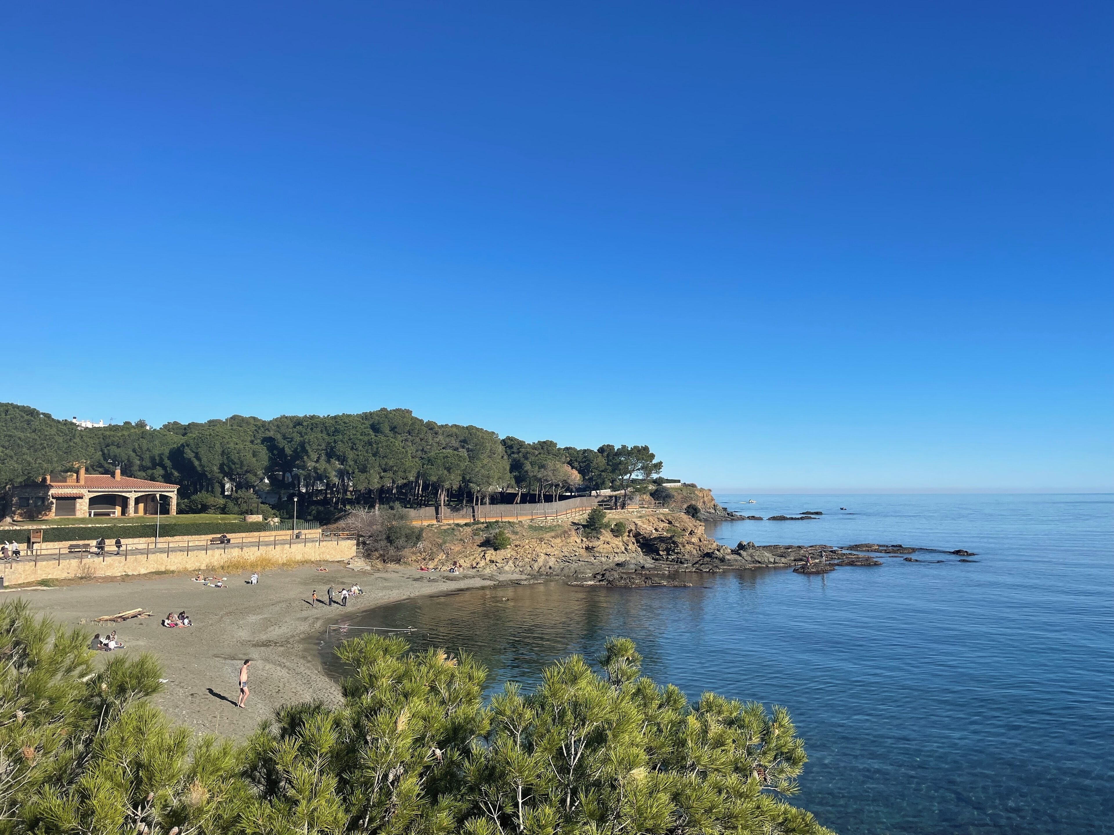 A coastal beach with a clear sky, rocky shoreline, and a building with a red roof in the background.