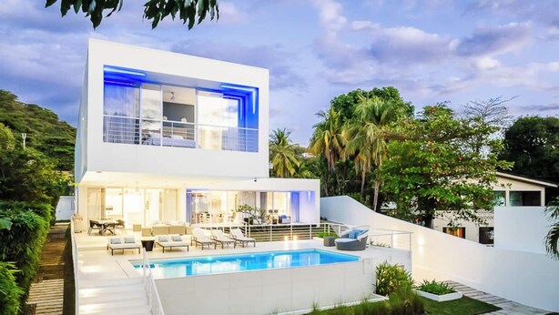 A modern two-story house with a swimming pool, outdoor seating, and lush greenery.