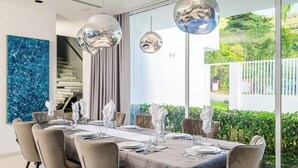 A dining area with a large table set for six, modern pendant lights, and a view of greenery outside.
