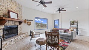 A living room with a stone fireplace, two ceiling fans, a large window with a view, a gray sofa, and a colorful rug. - Casa del Norte - Secluded Home in Del Norte Mtns (Alpine)