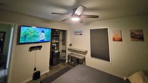 Un dormitorio con una cama, un televisor montado en la pared, un ventilador de techo, un microondas, un refrigerador y una ventana con persianas.