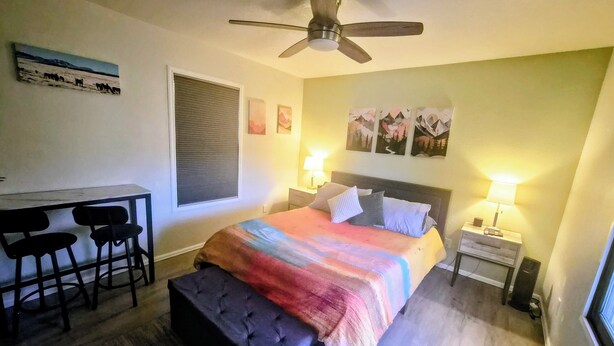 Un dormitorio con una cama, un escritorio con sillas, un ventilador de techo y obras de arte en las paredes.