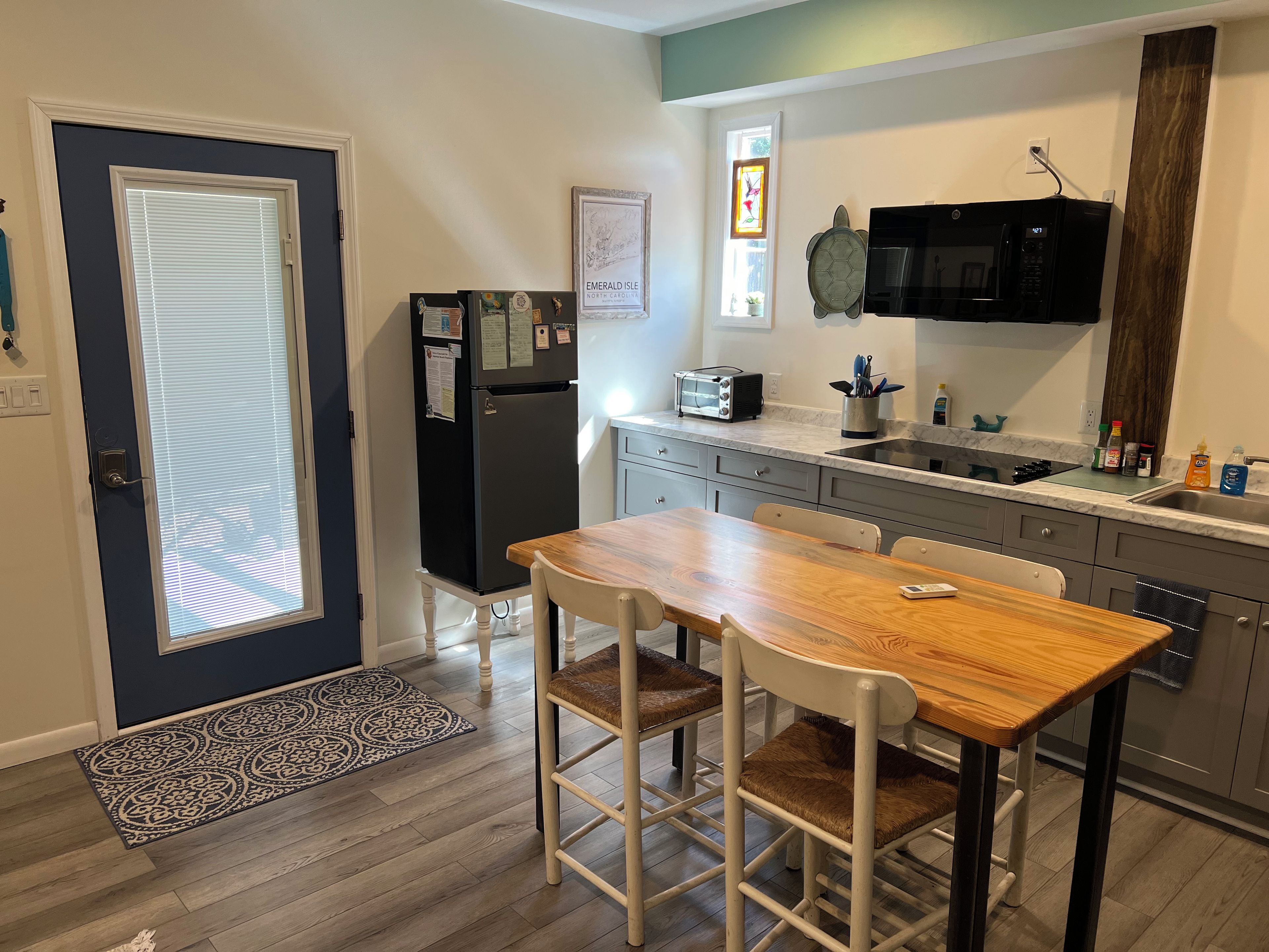 Een kleine keuken met een koelkast, magnetron en broodrooster. Er staat een eettafel met stoelen en een raam met luxaflex.