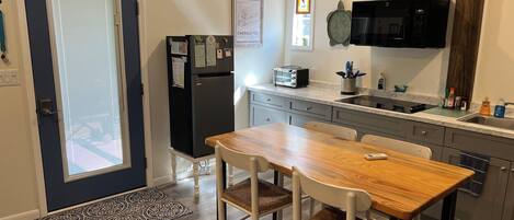 Een kleine keuken met een koelkast, magnetron en broodrooster. Er staat een eettafel met stoelen en een raam met luxaflex.