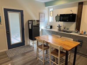 A compact kitchen with a refrigerator, microwave, and toaster. There is a dining table with chairs and a window with blinds.