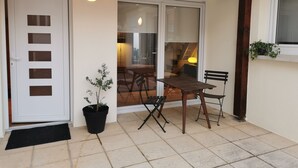 Een terras met een deur, een bloempot, een tafel met stoelen en een raam met een plant.