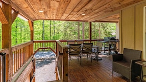 A covered outdoor deck with wooden flooring, a grill, and chairs. - Cabin close to town, private beach, parking & hot tub (Old Forge)