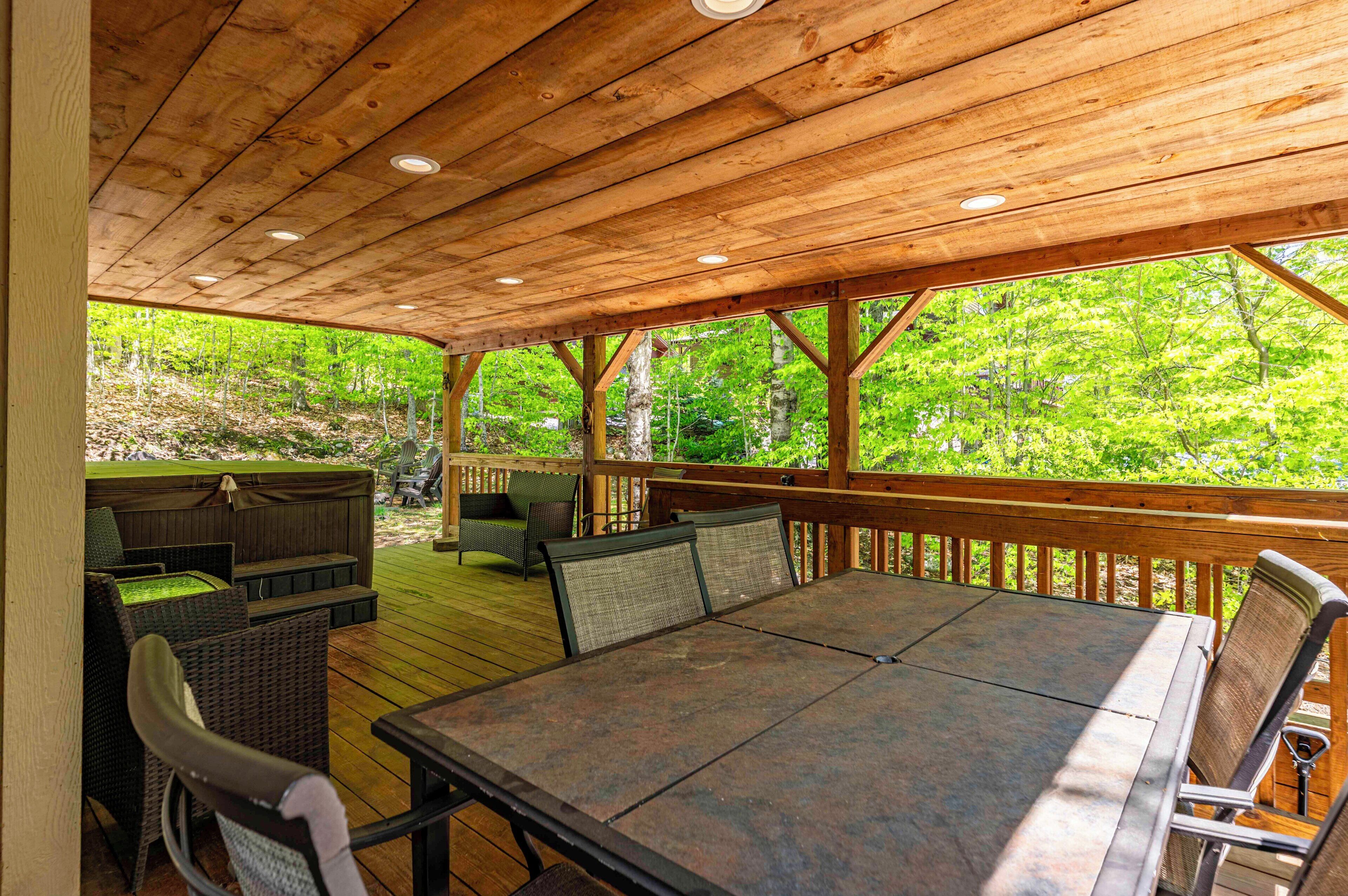 A covered outdoor deck with a table and chairs, surrounded by wooden railings and overlooking a forested area.