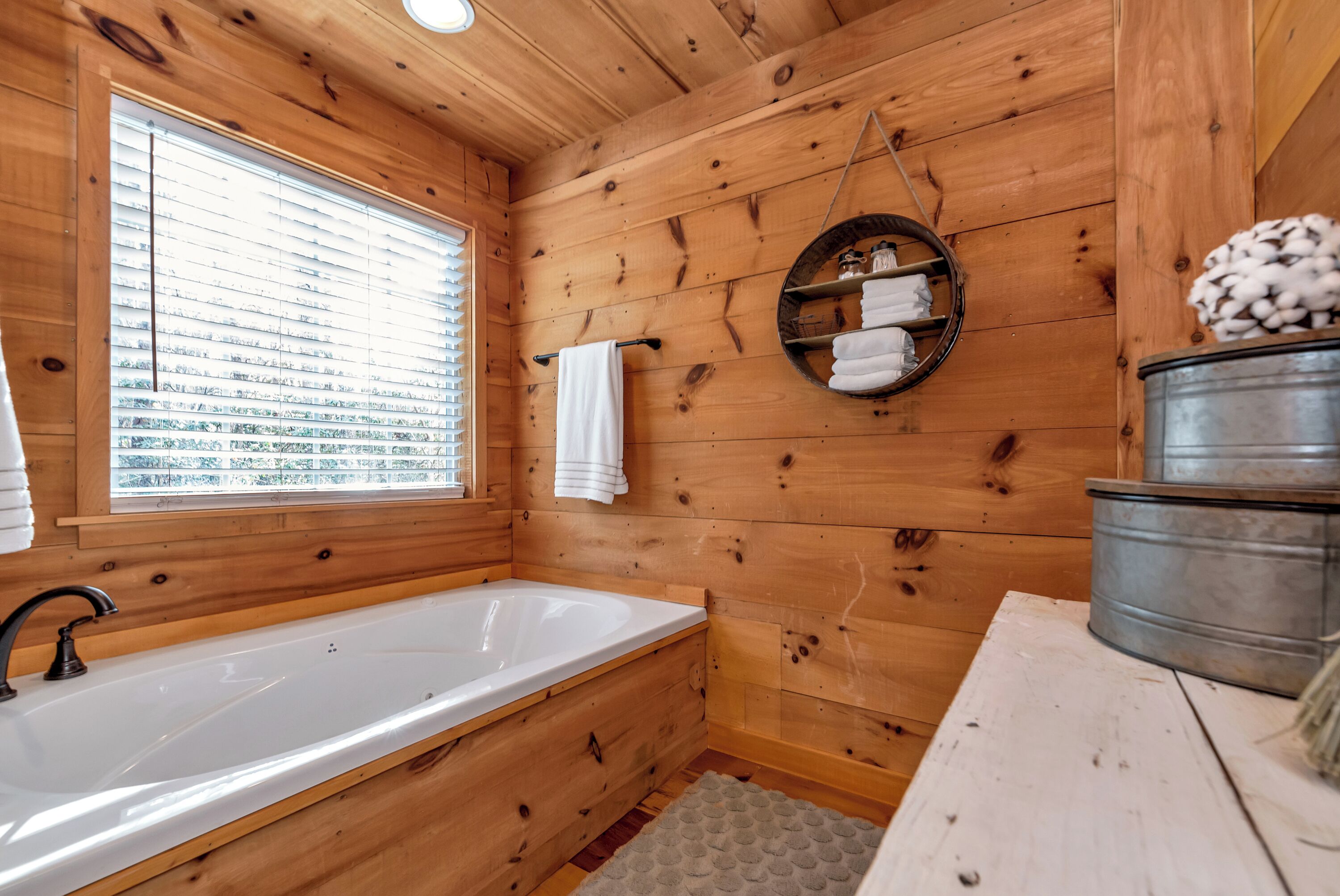 A wooden cabin bathroom with a large bathtub, a window with blinds, a towel rack, and a metal bucket.