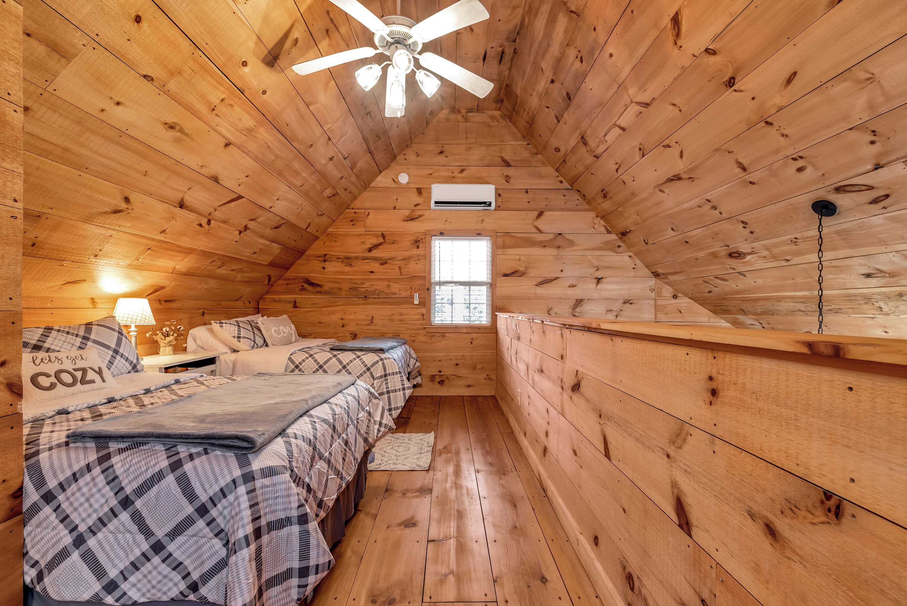 Ein gemütliches Schlafzimmer im Dachgeschoss mit Holzwänden, einer geneigten Decke, einem Deckenventilator, einem Fenster und einem Bett mit Karo-Bettwäsche.