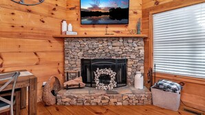 Interior acogedor de una cabaña con chimenea de piedra, paredes de madera, televisor montado y un cartel de bienvenida.