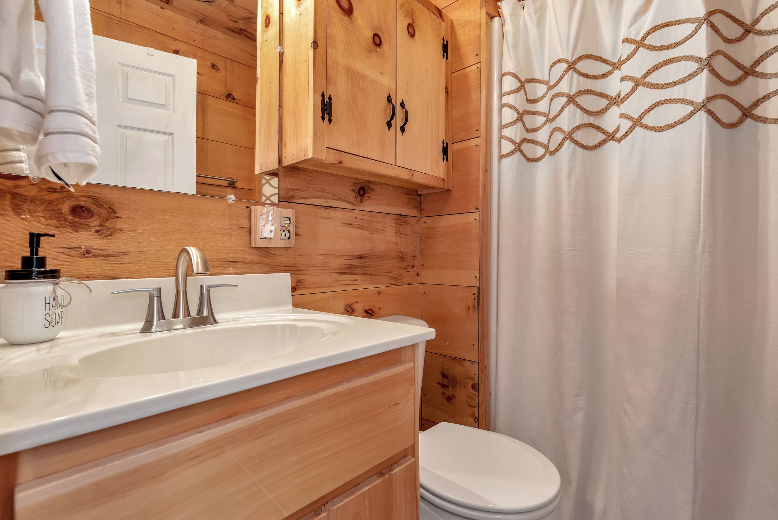 Un baño estilo cabaña de madera con un lavamanos blanco, un inodoro y una cortina de ducha con un diseño geométrico.