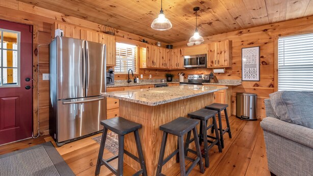 Interior de una cabaña de madera con cocina que incluye un refrigerador de acero inoxidable, un microondas y una isla de encimera de granito con taburetes de bar.