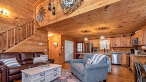 Interior acogedor de una cabaña con una escalera de madera, un sofá de cuero marrón, un sofá gris, un aparador blanco y una zona de cocina con armarios de madera y electrodomésticos de acero inoxidable.