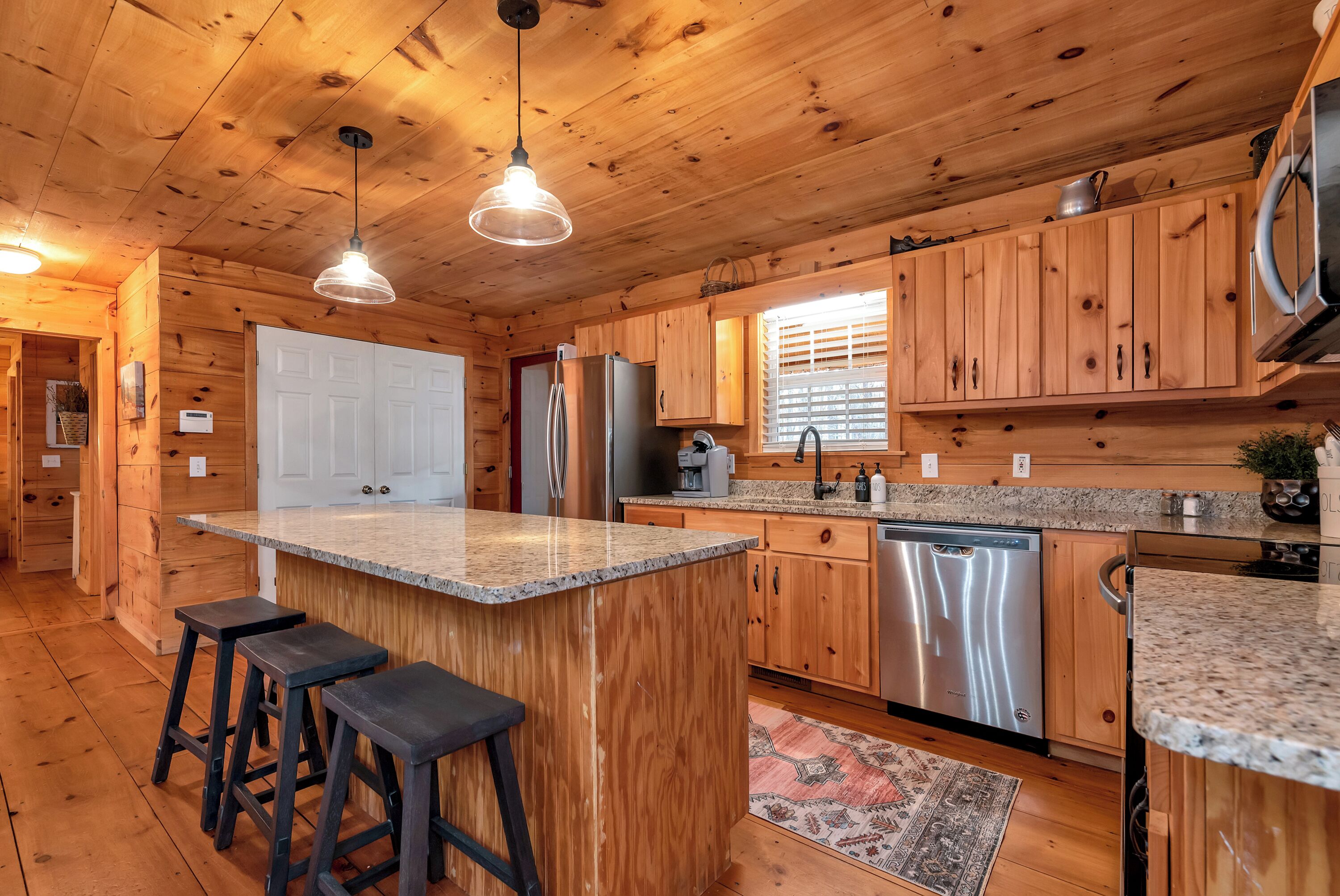 A wooden cabin kitchen with a central island, bar stools, stainless steel appliances, and a window with blinds.