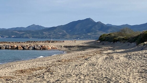 Een strand met een rotsachtige kustlijn, een pier en bergen op de achtergrond.