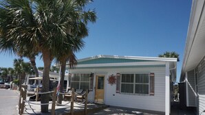 A small, single-story building with a white exterior and a green roof. It features a front door, a small porch with a bench, and a palm tree in the foreground. - Venture Out #41 Gulf Loop 2bd, 1ba WITH GOLF CART!!! (Panama City)