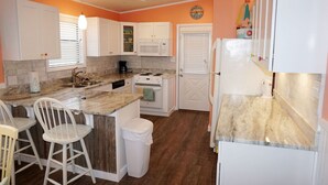 A compact kitchen with white cabinets, a microwave, a stove, and a sink. There are two white bar stools and a trash can. - Venture Out #41 Gulf Loop 2bd, 1ba WITH GOLF CART!!! (Panama City)