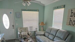 Una sala de estar con un sofá gris, un sillón estampado y una mesa de centro. Hay dos ventanas con persianas, un ventilador de techo y una puerta con ventana de vidrio.