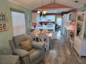 A living room with a sofa and chairs, a dining area with a table and chairs, and a kitchen with white cabinets and appliances.