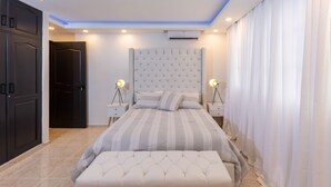 Quarto moderno com cabeceira estofada, cama grande com roupa de cama listrada, duas mesas de cabeceira com abajures e um banquinho aos pés da cama.