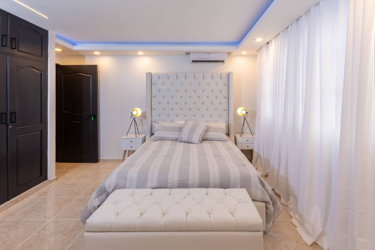 A modern bedroom with a tufted headboard, a large bed with striped bedding, two bedside tables with lamps, and a bench at the foot of the bed.