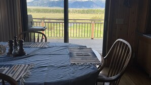A dining area with a table set for two, a view of mountains through large windows, and a wooden deck outside. - Spring Creek Guest House (Ennis)