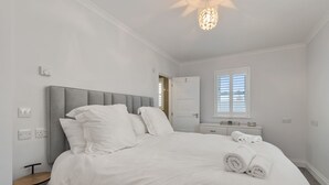 A bedroom with a large bed, a bedside table, a nightstand, a window with blinds, and a ceiling light fixture. - Cottage Mews - Seaside Cottage, Parking, Terrace (Tenby)