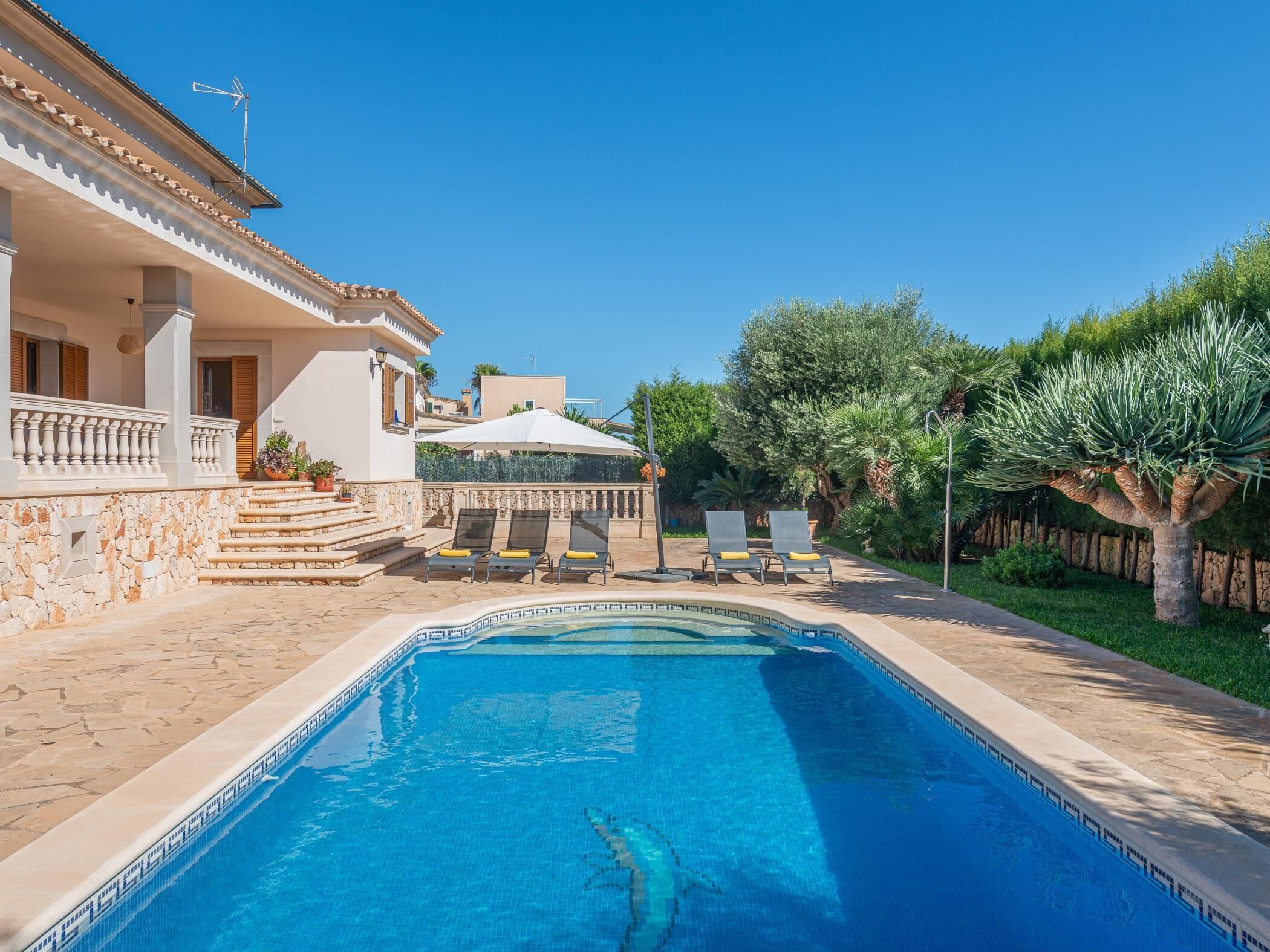 A swimming pool with a stone border, a covered patio with lounge chairs, and a stone staircase leading to a building.
