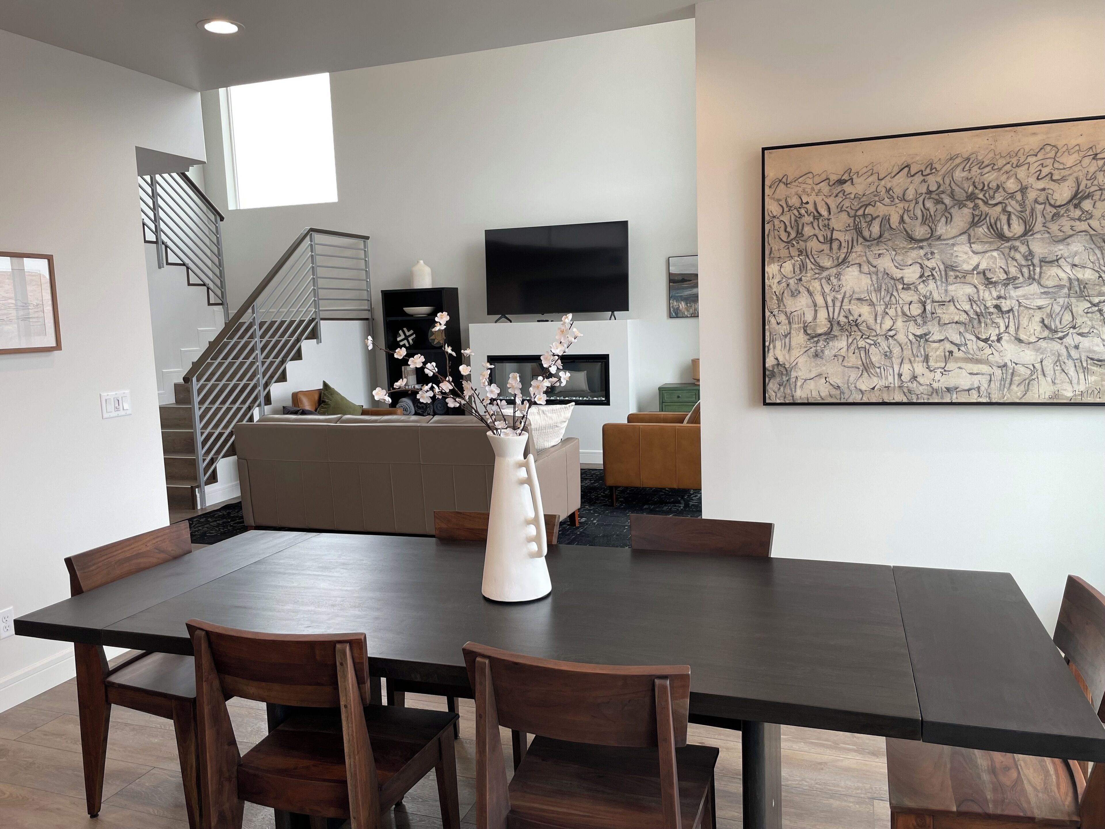 Een moderne eetruimte met een donker houten tafel en stoelen, een vaas met bloemen, een trap, een flatscreen televisie en een groot abstract schilderij aan de muur.