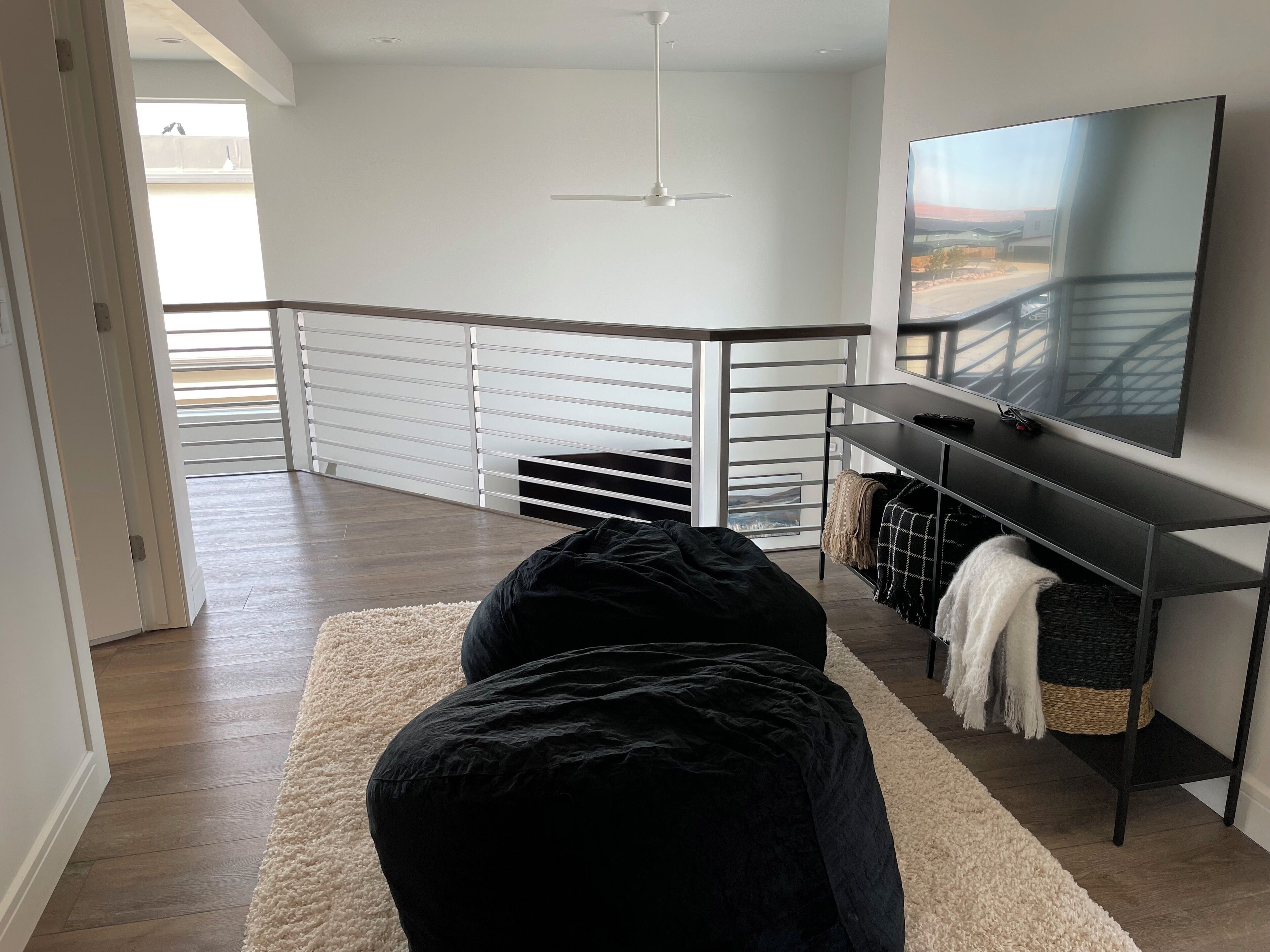 A modern living room with a black sofa, a white rug, a TV on a black stand, and a balcony with a railing.