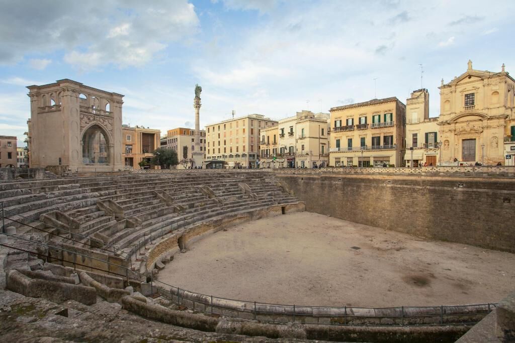 An ancient amphitheater with tiered seating and a central stage, surrounded by historic buildings.