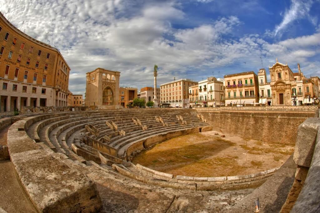 An ancient amphitheater with stone seating and a central stage, surrounded by historic buildings.