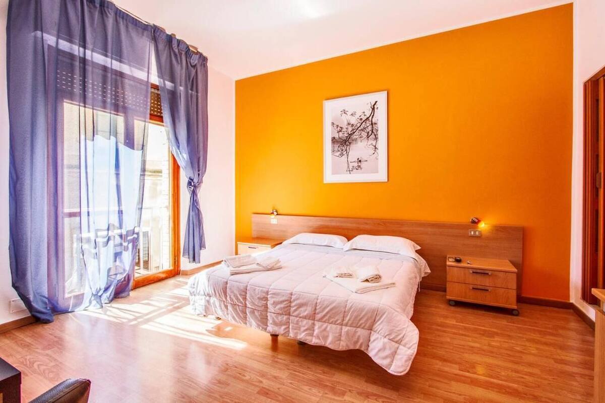Ein Schlafzimmer mit einem großen Bett, einem Nachttisch, einem Holz-Kopfstück, einem Fenster mit Vorhängen und einem gerahmten Kunstwerk an der Wand.