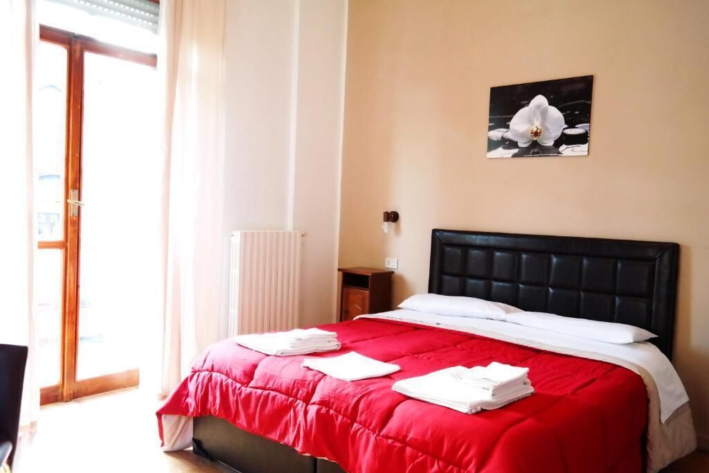 Ein Hotelzimmer mit Bett, rotem Überwurf, weißen Kissen, einem Blumenbild an der Wand und einem Fenster mit Vorhängen.