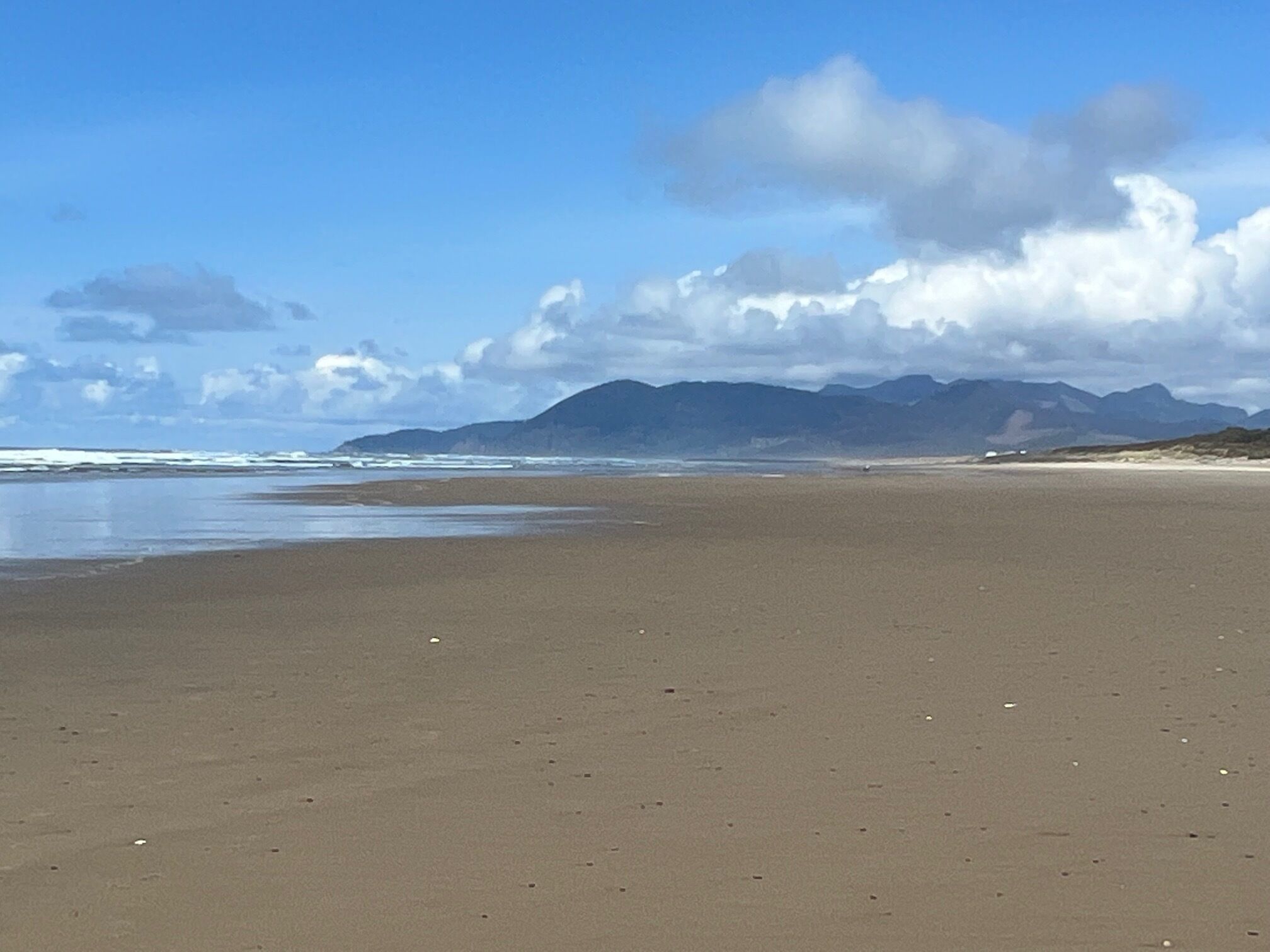 A wide sandy beach with gentle waves and distant mountains under a partly cloudy sky.