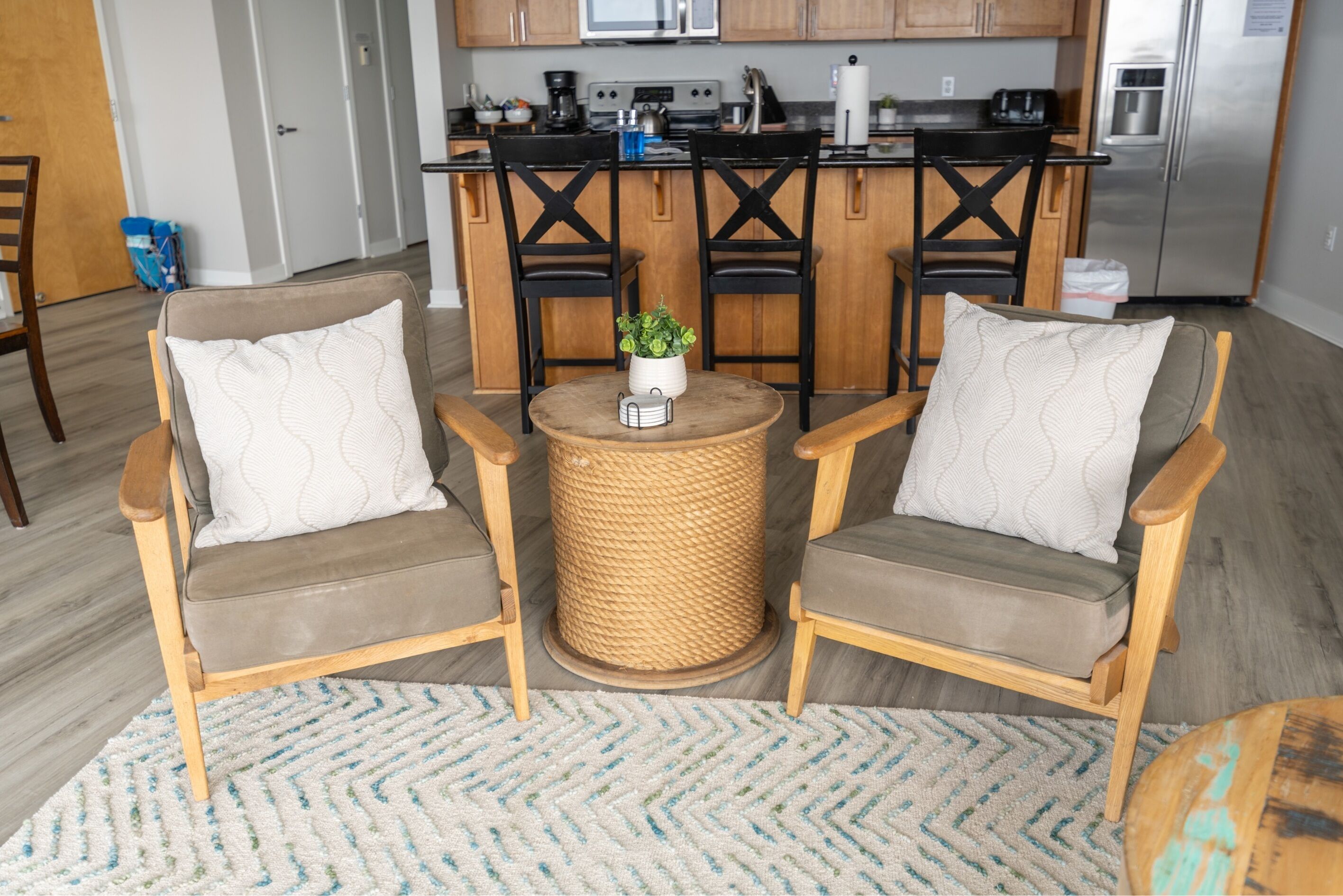 A cozy living room with two wooden chairs and a small round table, a kitchen in the background, and a wooden floor.