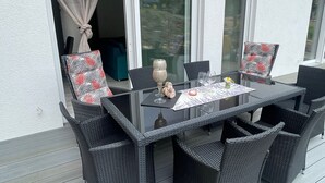 Un área de comedor al aire libre con una mesa de vidrio y sillas de mimbre, un florero y una pequeña mesa con una vela y un vaso.