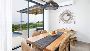 Un'area pranzo moderna con tavolo in legno, sedie e una ciotola di frutta. Si apre una grande finestra su una vista di spazi verdi e acqua.