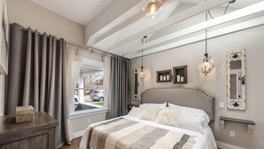 A bedroom with a large bed, a window with curtains, a bedside table, and a chandelier. - Agatha Stecker cottage - Historic Miner’s cottage (Louisville)